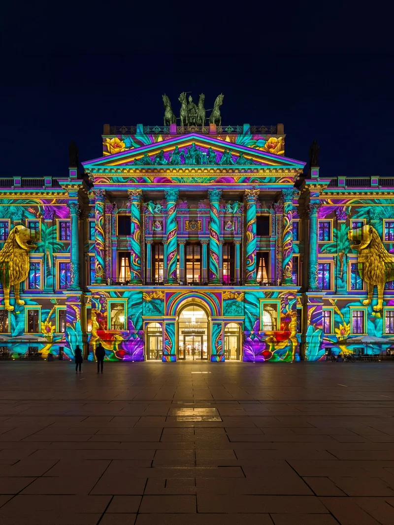 Night view of a historic building illuminated with vibrant, multicolored light projections, golden lion statues and floral patterns, with two silhouettes in the foreground.