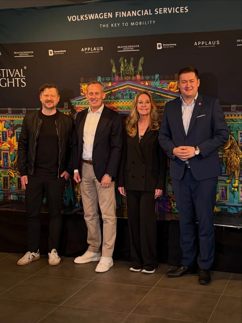 Four people pose before a dark backdrop displaying sponsor logos and a colorful projection of the illuminated building.