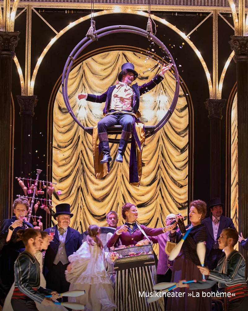 Stage with performer in top hat on a hoop, confetti falling, period-costumed audience; text 'Musiktheater »La bohème«' at bottom.