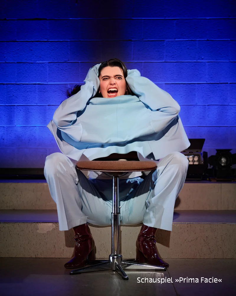 Actor in light blue outfit seated on a stool, hands over face, screaming, blue-lit brick wall; text 'Schauspiel »Prima Facie«' at bottom.
