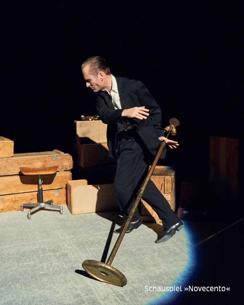 Man in dark suit leaning on a long pole with round base, wooden crates and stool on dim stage; text 'Schauspiel »Novecento«' at bottom.