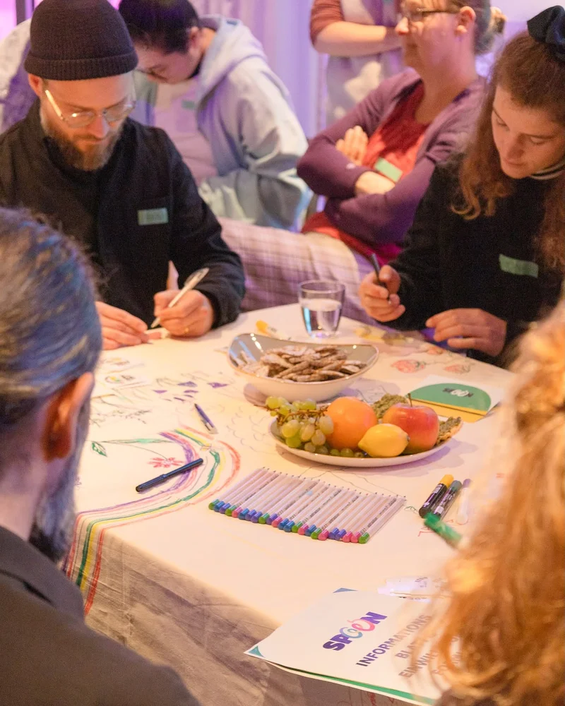 Participants drawing colorful rainbow patterns on a large sheet with markers and fruit nearby.